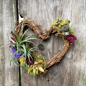 May include: A heart-shaped wreath made of brown twigs with a variety of succulents, air plants, and red berries. The wreath is hanging on a wooden fence.