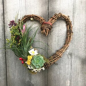 May include: A heart-shaped wreath made of natural twigs with a variety of air plants, succulents, and dried flowers. The wreath is hanging on a wooden wall.