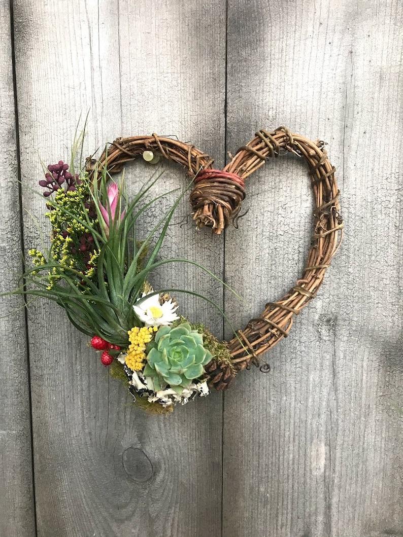 May include: A heart-shaped wreath made of natural twigs with a variety of air plants, succulents, and dried flowers. The wreath is hanging on a wooden wall.