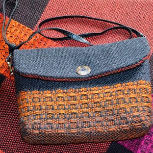 May include: A grey and orange woven crossbody bag with a black strap. The bag has a flap closure with a button and an orange woven pattern on the lower half. The edges are trimmed with red stitching.