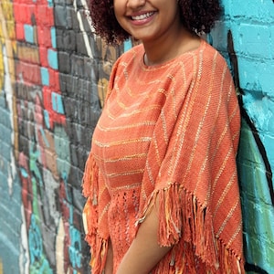 Può includere: Un poncho arancione con frange. Il poncho presenta un motivo a righe con dettagli dorati e bianchi. La persona indossa jeans azzurri e si appoggia a un colorato muro di graffiti.