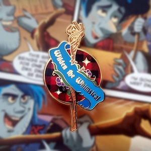 May include: A gold-colored enamel pin with a blue banner that reads "Wilder the Whimsical". The banner is wrapped around a brown stick with a gold-colored, twisted top. The background of the pin is a red and black plaid pattern with gold stars.