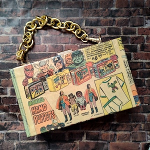 May include: A colorful comic book style clutch purse with a gold chain strap. The front of the purse features a variety of Marvel characters, including Spider-Man, Superman, and the Hulk. The text on the purse reads "Marvel Hand Puppets".