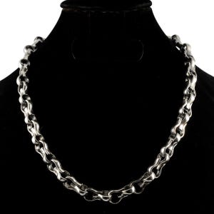 Puede incluir: Un collar de cadena en tono plateado que presenta eslabones redondeados gruesos entrelazados con un acabado metálico pulido. El collar se muestra en forma circular, destacando la construcción uniforme y resistente de los eslabones metálicos.