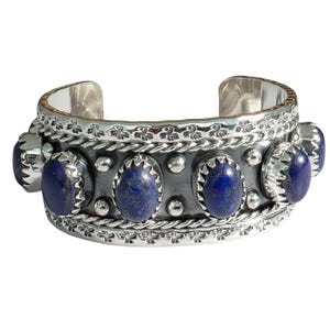 Puede incluir: Brazalete de plata con gemas ovaladas de color azul oscuro. El brazalete tiene un diseño decorativo con un borde en forma de cuerda y pequeños detalles plateados. Las gemas están engastadas en un engaste de metal oscuro.