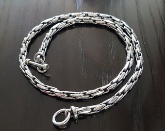 Puede incluir: Un collar de cadena de plata con cierre de palanca. La cadena está hecha de pequeños eslabones planos que se entrelazan en un patrón.