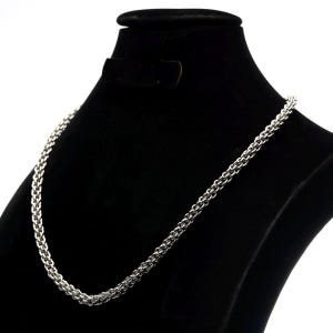 May include: A silver-toned chain necklace featuring a dense, woven link pattern. The chain is displayed on a black velvet-textured bust, highlighting the metallic sheen and the intricate, repeating structure of the interconnected metal links.