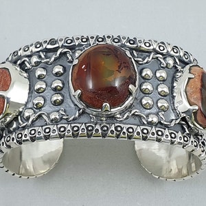 Puede incluir: Un brazalete de plata con tres piedras ovaladas de color rojo y marrón engastadas en el centro. El brazalete tiene un diseño detallado con cuentas de plata y un patrón de cadena.