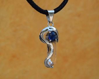Puede incluir: Un collar de plata con un colgante de cristal de cuarzo transparente y una piedra preciosa azul. El collar está en un cordón negro.