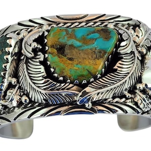 Puede incluir: Una pulsera de plata ancha tipo brazalete con tres grandes piedras de turquesa irregulares con matriz marrón. La superficie de plata está intrincadamente grabada con motivos de plumas detallados y volutas decorativas alrededor de la piedra central.