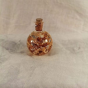 Puede incluir: Una pequeña botella redonda de vidrio con un tapón de corcho. La botella está decorada con pan de oro y tiene una superficie texturizada.