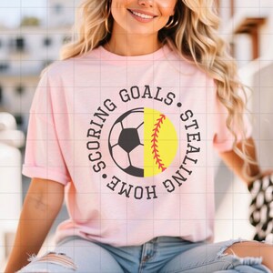 May include: Pink t-shirt with a black circle graphic featuring a half soccer ball and half softball with the text "Scoring Goals. Stealing Home."