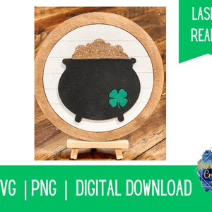 Puede incluir: Un letrero redondo de madera con fondo blanco y una olla de oro negra con un trébol verde. El letrero está en un caballete de madera. El texto "Laser Ready" está en una pancarta verde en la parte superior de la imagen.