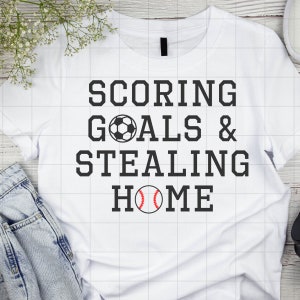 May include: White t-shirt with black text that reads "SCORING GOALS & STEALING HOME". A soccer ball and a baseball are included in the text.