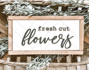 May include: A wooden sign with a white background and a black inscription that reads "fresh cut flowers". The sign is framed in a natural wood frame and is surrounded by green foliage.