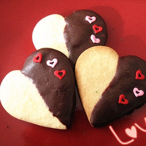 May include: Three heart-shaped cookies decorated with pink and red sprinkles. The cookies are half white and half dark chocolate. The cookies are on a red background with the word "Love" written in white.