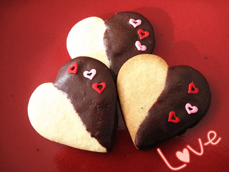 May include: Three heart-shaped cookies decorated with pink and red sprinkles. The cookies are half white and half dark chocolate. The cookies are on a red background with the word "Love" written in white.