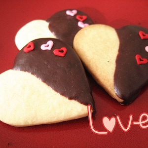 May include: Three heart-shaped cookies decorated with pink and red sprinkles. The cookies are half white and half dark chocolate. The cookies are on a red surface with the word "love" written in pink.