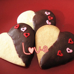 May include: Three heart-shaped cookies decorated with red and pink sprinkles. The cookies are half white and half dark chocolate. The word "Love" is written in pink and white on the cookies.