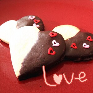 May include: Two heart-shaped cookies decorated with pink and red sprinkles. The cookies are half white and half dark chocolate. The cookies are on a red plate with the word "Love" written in pink.