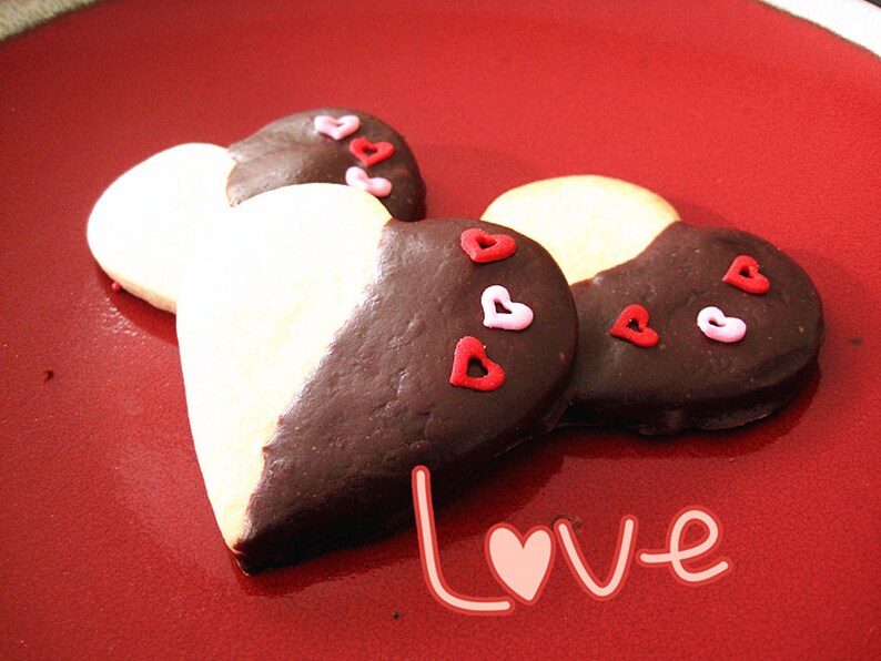 May include: Two heart-shaped cookies decorated with pink and red sprinkles. The cookies are half white and half dark chocolate. The cookies are on a red plate with the word "Love" written in pink.