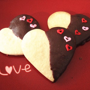 May include: Three heart-shaped cookies decorated with red and pink sprinkles. The cookies are half white and half dark chocolate. The cookies are on a red surface with the word "Love" written in pink.