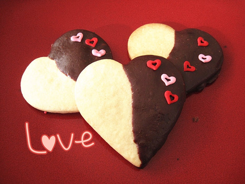 May include: Three heart-shaped cookies decorated with red and pink sprinkles. The cookies are half white and half dark chocolate. The cookies are on a red surface with the word "Love" written in pink.