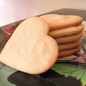 May include: A stack of heart-shaped sugar cookies on a floral patterned surface. The cookies are pale yellow and have a slightly rough texture.