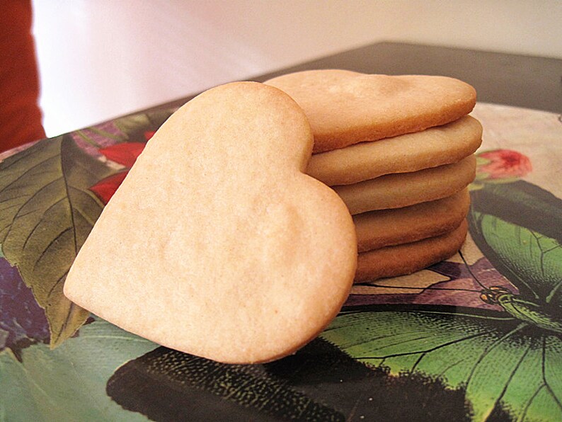 May include: A stack of heart-shaped sugar cookies on a floral patterned surface. The cookies are pale yellow and have a slightly rough texture.