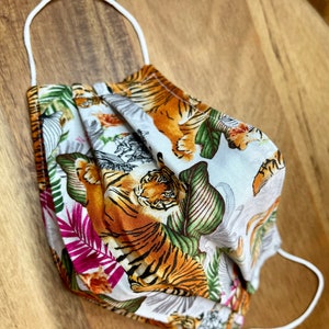 May include: A white face mask with a repeating pattern of orange and black tigers and green leaves. The mask has white elastic straps.