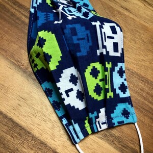May include: A blue face mask with a white elastic band. The mask has a pixelated pattern of skulls, bones, and geometric shapes in shades of blue, green, and white.