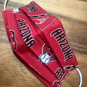 May include: Red face mask with a white elastic band. The mask has a repeating pattern of a cartoon snake with a baseball in its mouth, the word "Arizona" and a stylized "A" with a diamond inside.