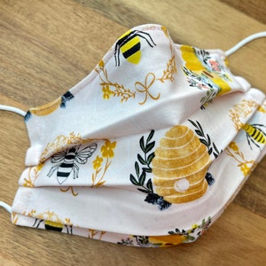 May include: A pink face mask with a bee and honeycomb pattern. The mask has white elastic straps.