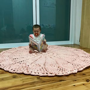 May include: A pink crocheted round rug with a delicate lace pattern. A baby is sitting on the rug, reading a book.