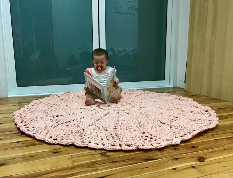 May include: A pink crocheted round rug with a delicate lace pattern. A baby is sitting on the rug, reading a book.