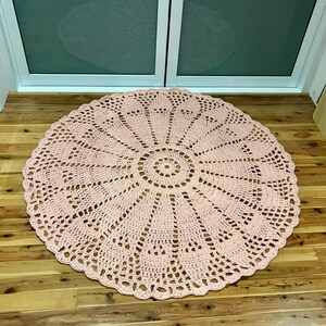 May include: A large, round, pink crocheted rug with a delicate, intricate pattern. The rug has a circular design with a central circle and radiating spokes.