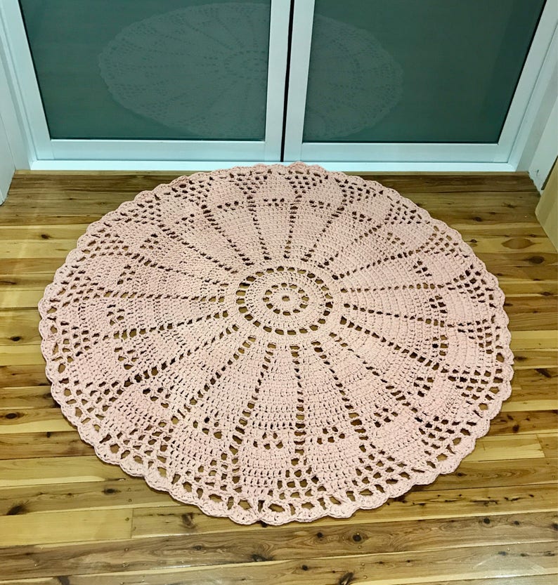 May include: A large, round, pink crocheted rug with a delicate, intricate pattern. The rug has a circular design with a central circle and radiating spokes.