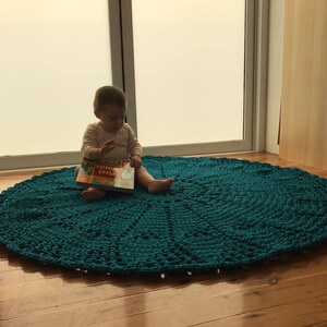 May include: A teal crocheted round rug with a child sitting on it reading a book titled "First Words".