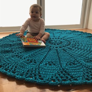 May include: A baby sits on a teal blue crocheted rug, holding a book titled "First Words".