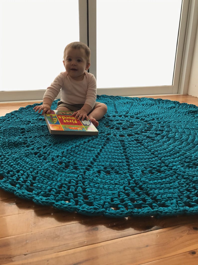 May include: A baby sits on a teal blue crocheted rug, holding a book titled "First Words".