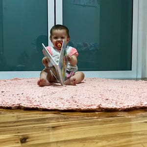 May include: A baby sitting on a pink crocheted rug, holding a colorful book and wearing a pacifier.