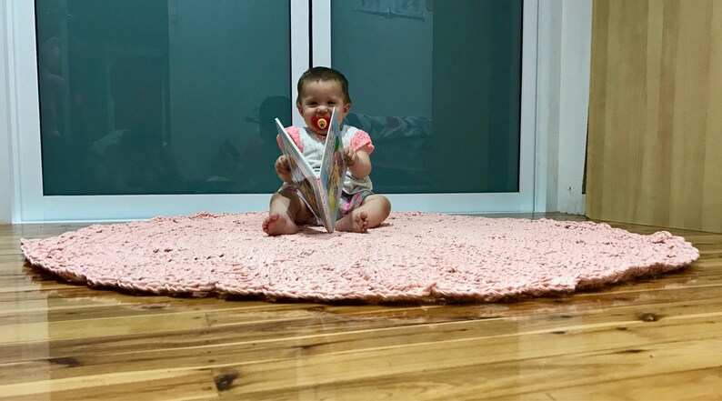 May include: A baby sitting on a pink crocheted rug, holding a colorful book and wearing a pacifier.