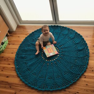 May include: A teal crocheted round rug with a baby sitting on it, holding a book titled "First Words".