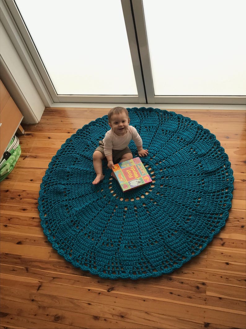 May include: A teal crocheted round rug with a baby sitting on it, holding a book titled "First Words".