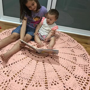 May include: Two children, one sitting on the other's lap, read a book on a pink crocheted round rug. The rug has a lacy design.