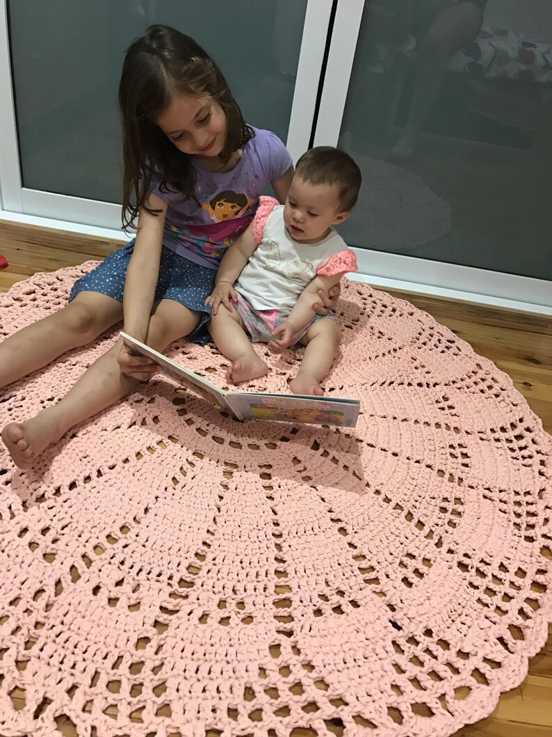 May include: Two children, one sitting on the other's lap, read a book on a pink crocheted round rug. The rug has a lacy design.