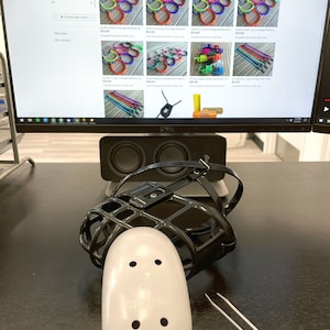 May include: A black dog muzzle with a white padded nose guard and two white plastic cable ties. The muzzle is made of metal and has a black strap. The muzzle is on a dark brown surface.