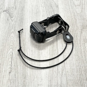 Könnte beinhalten: Schwarzes Hundetraining-Halsband mit schwarzem Empfänger und schwarzem Riemen. Das Halsband ist an einem schwarzen Gummiband befestigt.