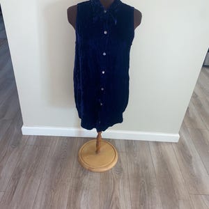 Könnte beinhalten: Ein ärmelloses, dunkelblaues Samtkleid mit Knopfleiste wird auf einer Schaufensterpuppe präsentiert. Das Kleid hat einen Kragen und eine leicht ausgestellte Form. Die Schaufensterpuppe steht auf einem Holzständer.