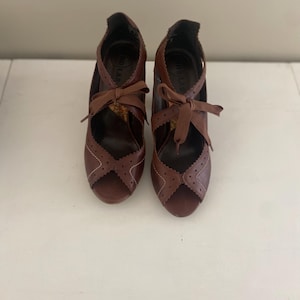 May include: A pair of brown leather peep-toe heels with decorative cutouts and ribbon ties. The shoes have a high heel and a scalloped edge design. The shoes are displayed on a white surface.